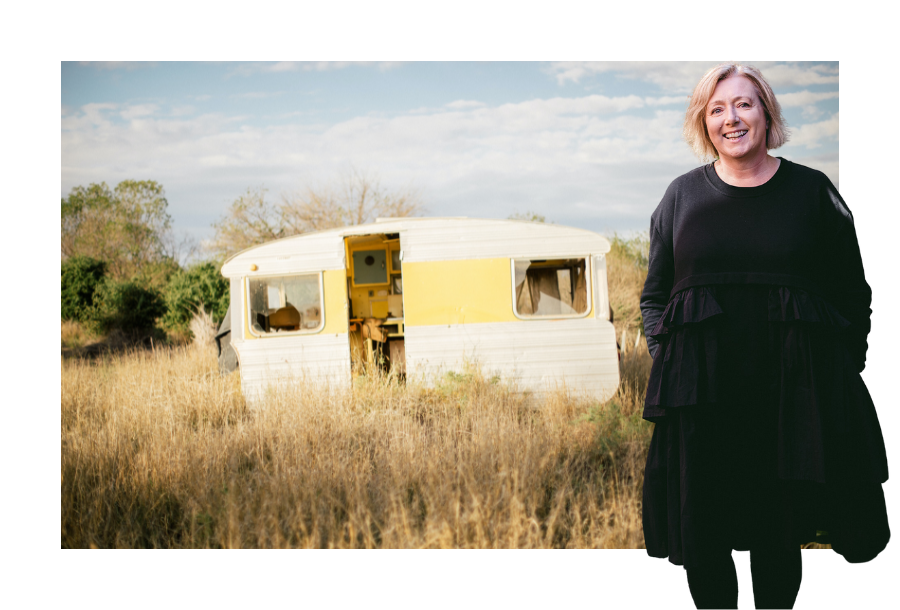 Pictured, a yellow and white coloured caravan in a deserted paddock with overgrown dead grass and broken windows. In the forefront stands Krys Canny-Smith wearing a back dress with her hands in her pockets and black stockings.