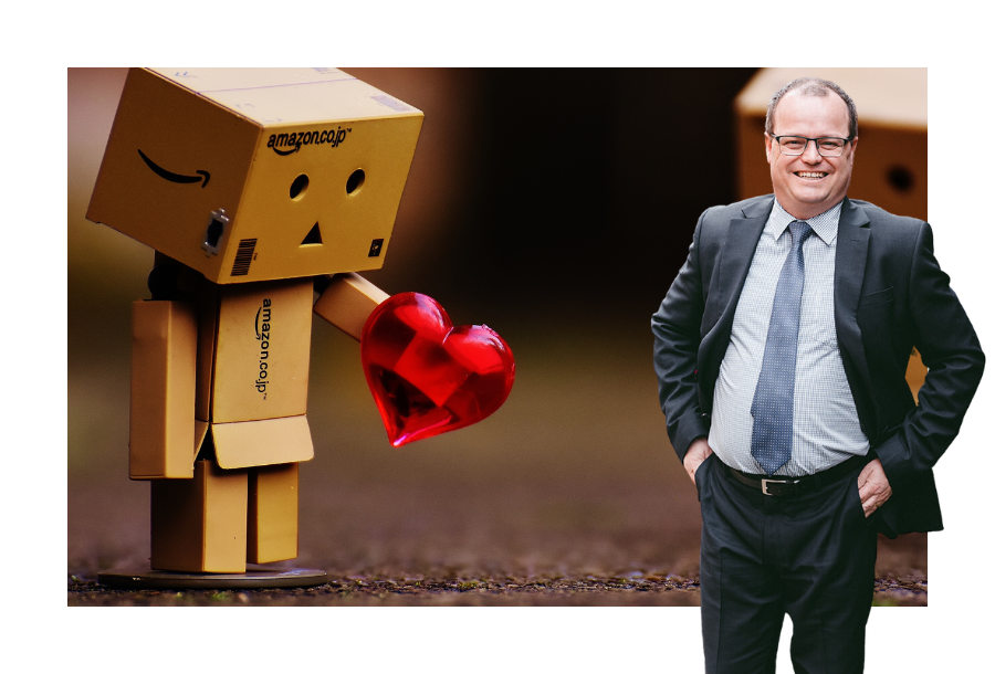 Pictured, a cardboard box looking sad holding a 3D red heart. On the right hand side, stands Adam Wightman with his hands in his pockets of his dark grey suit.