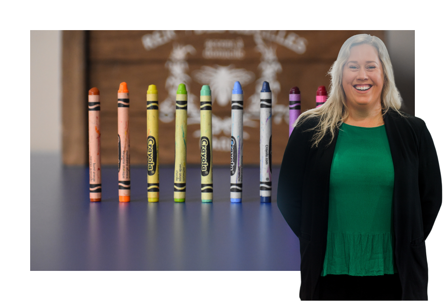 Pictured, Katie Greig with a big smile and her hands behind her back, wearing a black blazer and a eep green coloured top. Behind her is a stock picture of different coloured Crayola crayons all standing upright with a small space between them.