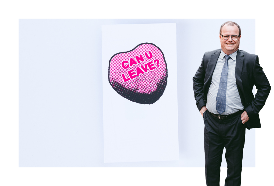 Pictured, Adam Wightman with his hands on his hips, his suit jacket undone and a big smile on his face. Behind Adam is a picture of a hand drawn 3D lolly that says "Can U Leave?" in the shape of a love heart.