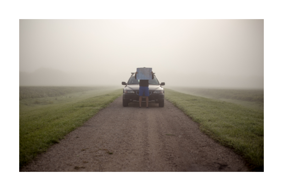 Pictured, a misty sky and open green fields. In the centre is a road and a car parked in the distance. There is a person sitting on the hood of the car holding a map between their hands hiding their face.