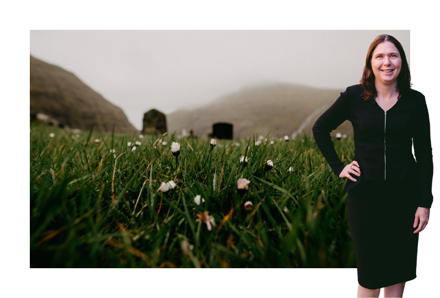 Pictured, Karlene Wightman wearing a long sleeve black dress standing with on hand on her hip and a big smile. In the background is a stock picture of a misty field, with small white flowers in the foreground and headstones in the background.