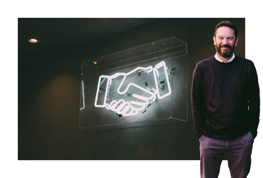 Pictured, Danny Grigg with his hands in his pockets wearing a long sleeve black jumper. Standing in front of a picture that is a black background and two white neon lit hands doing a hand shake.