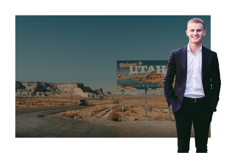 Pictured, Jayden Scott wearing a dark black coloured suit and his hands in his pockets. Standing in front of a picture of a billboard that says "UTAH life elevated" which sits on an intersection of a deserted road and mountains in the background.