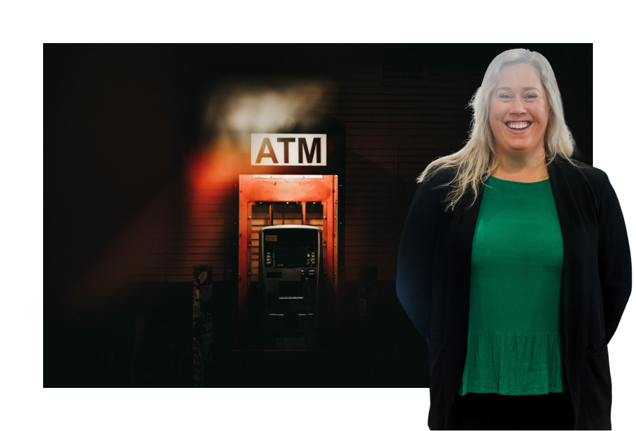 Pictured, Katie Greig with a big smile on her face and her hands behind her back. Wearing a dark green top and a black blazer, she is standing in front of a picture with a dark background and a light shinning on an orange coloured ATM, with the sign "ATM"