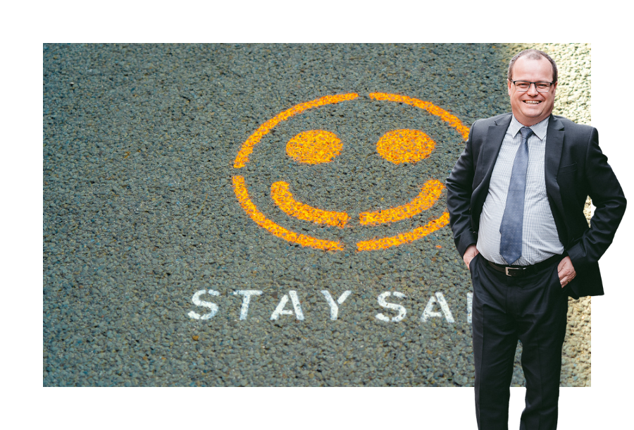 Pictured, Adam Wightman with his hands in his suit pant pockets and a big smile on his face. Standing in front of a bitumen walk way, with the words printed in white paint "stay safe" and a picture of a yellow smiley face.