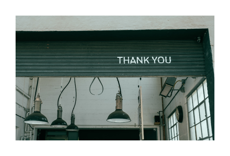 Pictured, a high view of the inside of a workshop, you can see the white brick work with lots of different hanging lights coming from the ceiling. On the outside, a dark green roller door is 3/4 of the way open with the words painted in white "thank you"