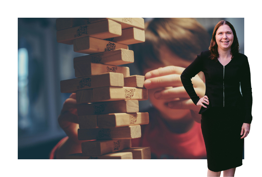Pictured, Karlene Wightman wearing a long sleeve black dress and one hand on her hip. With her brown hair touching her shoulders she also has a big smile on her face. In the background is a muted tones picture of a little child playing a game of Jenga by himself.