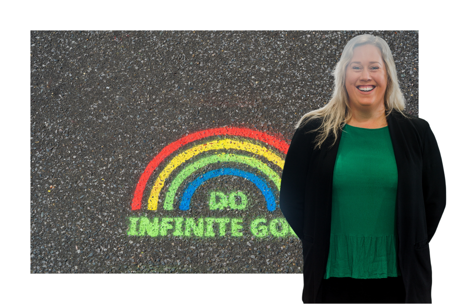 Pictured, Katie Greig standing with her hands behind her back and a big smile on her face. In the background is a picture drawn in bright coloured red, yellow, green and blue chalk on the pavement. In the shape of a rainbow with the words "DO INFINITE GOOD"