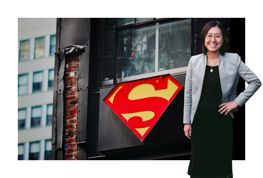 Pictured, Helen Yau with a big smile on her face, one hand on her hip wearing a black dress and a teal-coloured blazer. In the background is a street sign with the superman logo in bright red and yellow against a black wall.