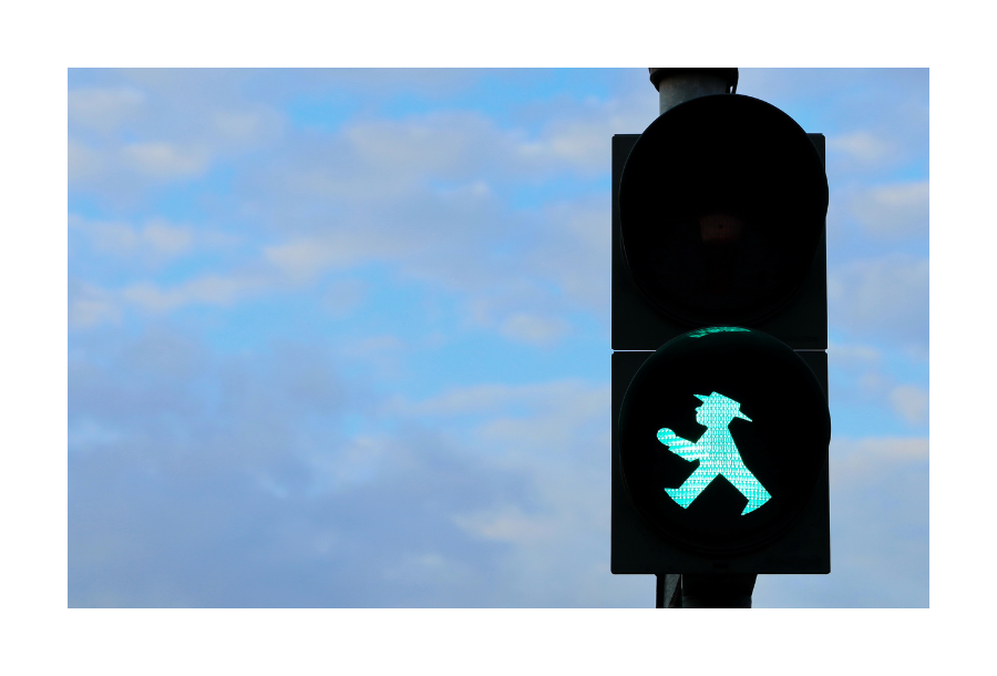 Pictured, a blue sky with light grey clouds in the background, in the forefront is a traffic light with the green walking man illuminated.