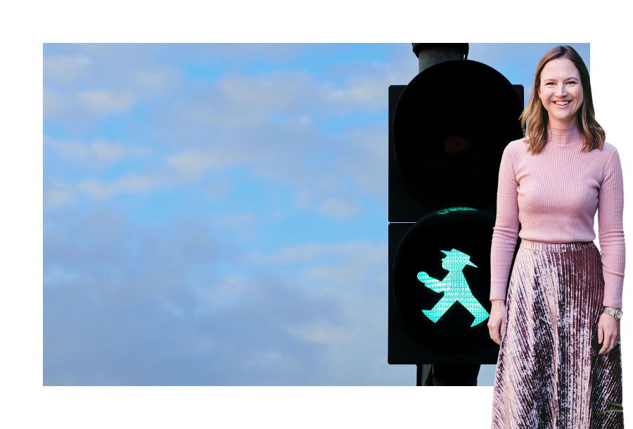 Pictured, Gabrielle Andersen wearing a pink velvet skirt and a long sleep pink top. In the background is a picture of a green walking man on a street crossing sign, against a beautiful blue cloudy sky.