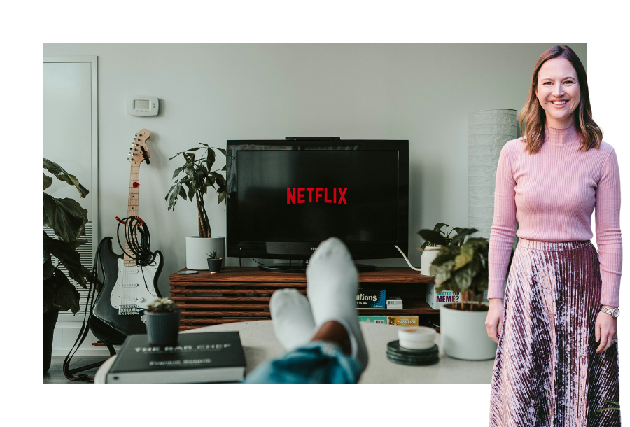 Gabrielle Andersen stands wearing a pink velvet skirt and a long sleep pink skivvy. In the background, you can see a pair of feet wearing white socks and legs crossed on a coffee table with a television turned on and the words "NETLIX" on the screen.