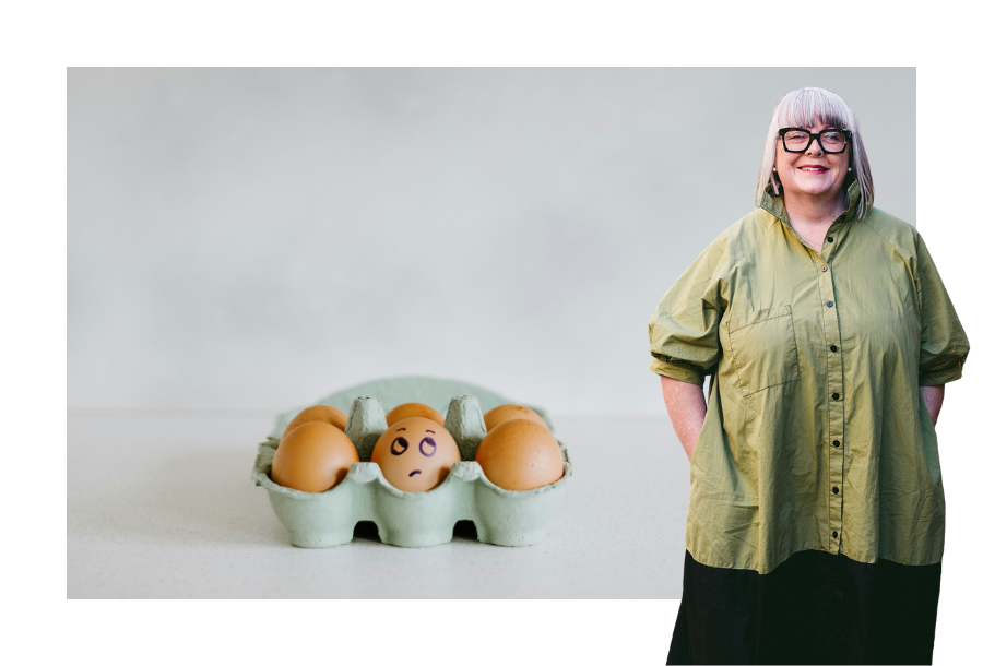 Pictured, Anthea Taylor with her hands in her pockets and a big smile on her face. Wearing an oversized olive green coloured top and a black skirt. In the background against a white bench and wall is a 6-way egg carton with 6 eggs, one has a drawing of eyes and eyebrows making the face of someone rolling their eyes.