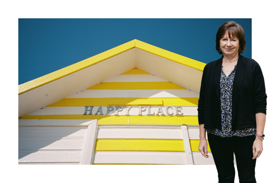 Pictured: Nancy Robertson standing smiling with her hands by her side, wearing a floral top with black pants and black cardigan. Behind her is an image of a yellow and white house roof with a sign on the wall that says "Happy place".