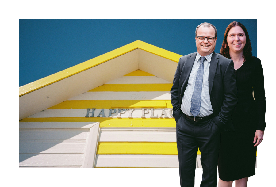Pictured, Adam Wightman and Karlene Wightman standing in front of a picture of a brightly painted beach shake - seeing only the pointed roof, it's bright yellow and white stripes with the words "HAPPY PLACE" in the centre.