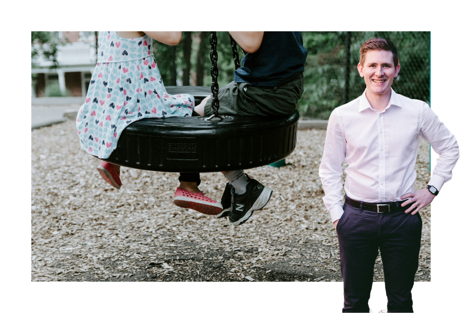 Pictured, Chris Graham with one hand on his hip and a big smile. In the background, there are two kids on a tyre swinging in a park.