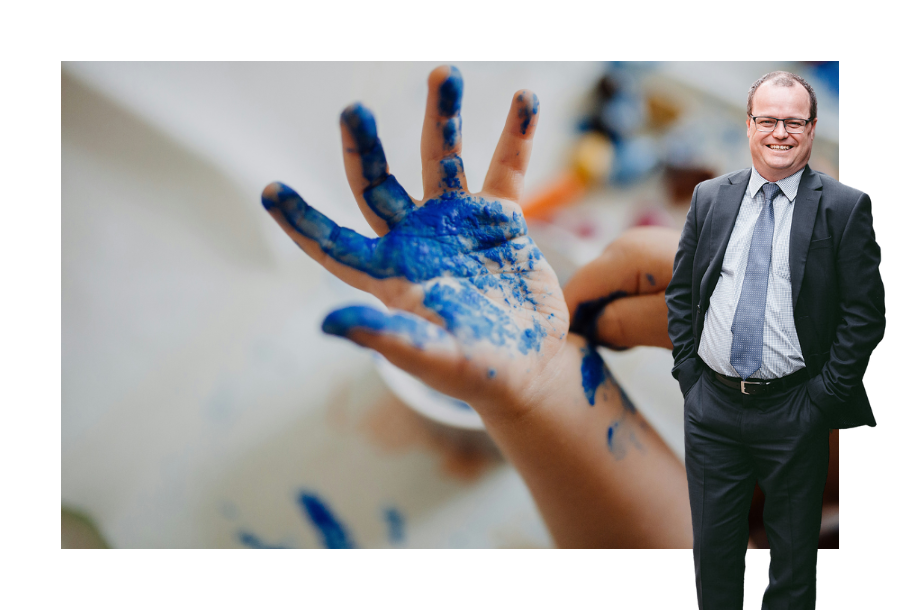 Pictured, Adam Wightman standing in a dark grey suit with his hands in his pockets and a big smile on his face. In the background is a picture of a child's hand and fingers are wriggling, that is covered in dark blue paint.