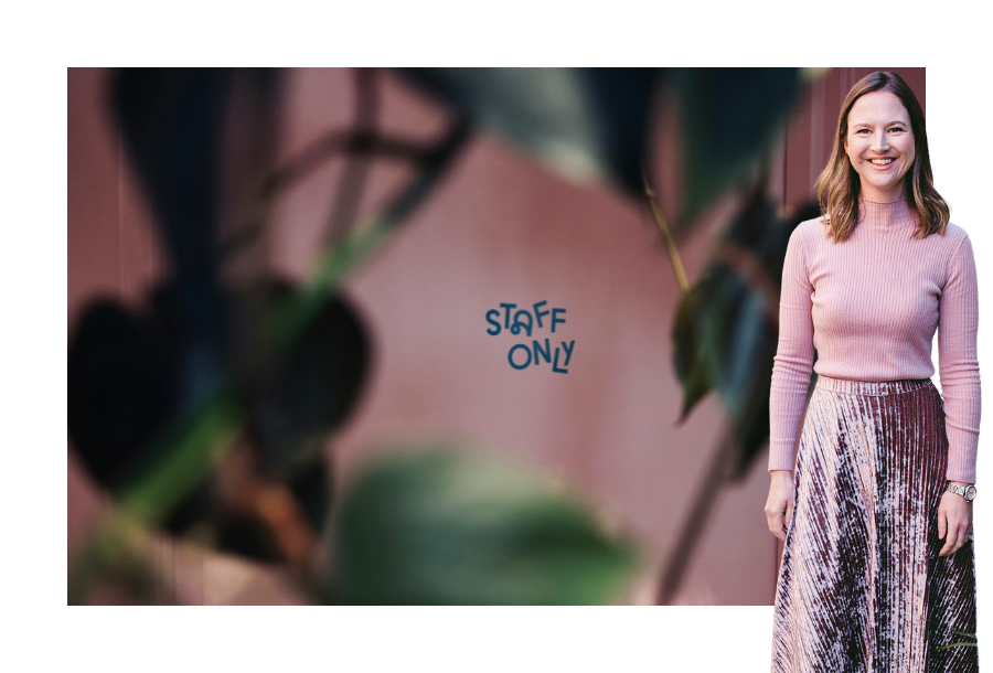 Pictured: Gabrielle Andersen, Head of Canny Insight. Standing and smiling wearing a pink sweater and velvet pink long skirt. Behind her is a photo of a pink door taken through some indoor plants, written on the door are the words "Staff Only".