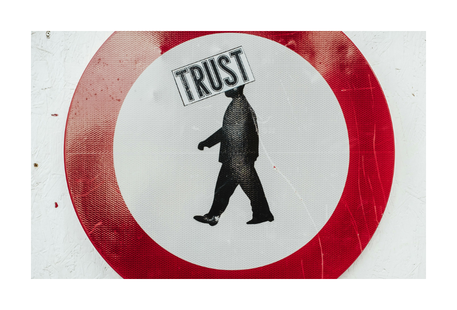 Pictured, a white sign with a thick red circle boarder, inside of the red circle is a black and white stencil picture of a person walking - where the head should be is the word "TRUST" in capital letters.