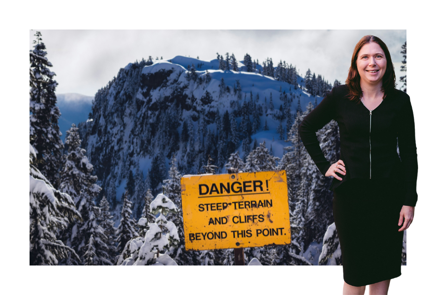 Pictured, Karlene Wightman with one hand on her hip and a big smile on her face. In the background is a picture of a snowy cliff with a sign that reads "DANGER! Steep terrain and cliffs beyond this point.