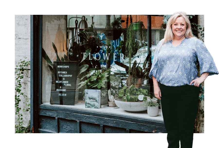 Pictured; Michelle Flowers standing in a blue shirt and black pants with her hands on her hips smiling. Behind her is a photo taken above a register and eftpos machine.