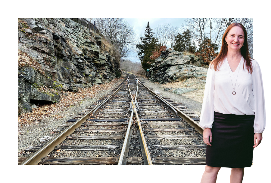 Pictured; Samantha Butcher, standing smiling in a white long sleeve top and a black skirt, behind her is an image of two railway tracks converging into one track.
