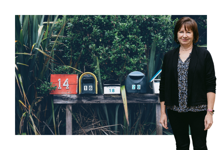 Pictured: Nancy Robertson standing smiling with her hands by her side, wearing a floral top with black pants and black cardigan. Behind her is an image of colourful mailboxes lined up on a post backed by bushes.