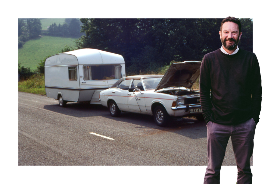 Pictured; Senior Accountant, Danny Grigg, standing smiling with his hands in his pockets wearing a black sweater and brown trousers, behind him is a photo of an car broken down while towing a caravan with a green hillside in the background.