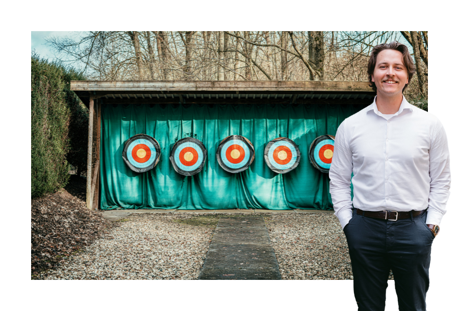 Pictured, Zachary Henderson standing smiling in white business button up and navy-blue trousers. Behind him is a photo of targets setup against a wall with a blue curtain, outside in the woods.