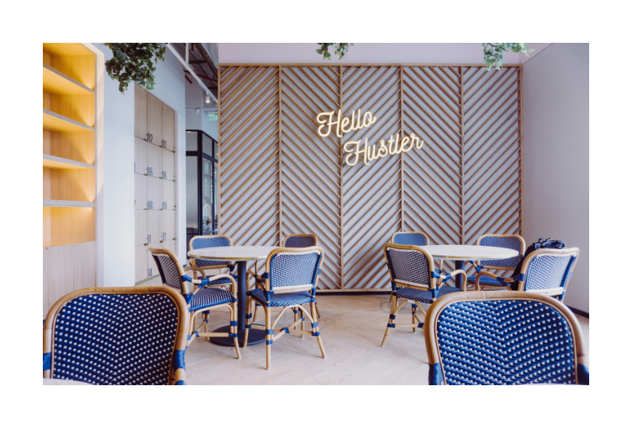 Pictured, a beautiful working space, clean tiled floor and a wooden slated feature wall with a neon sign lit up saying "hello hustler" there are four working tables spaced around with blue and white rattan style chairs.