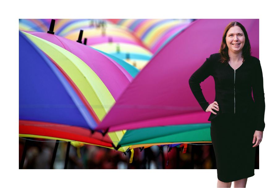 Pictured, Karlene Wightman wearing a black long sleeve dress, one hand on her hip and a big smile on her face. In the background is a picture of many umbrellas all opened and of different colours.