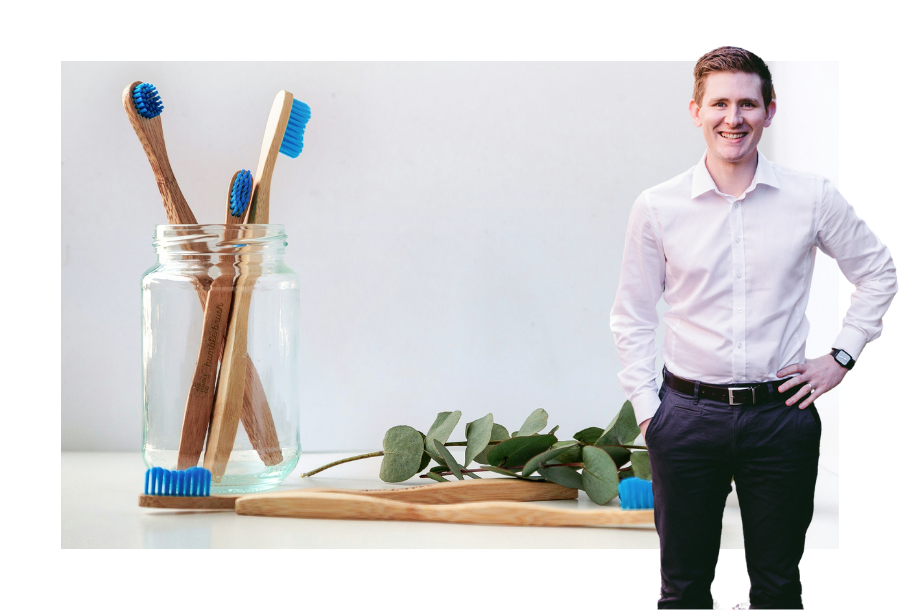 Pictured, Chris Graham with one hand on his hip and a big smile. In the background there is an image of wooden toothbrushes with a sprig of leaves laid down next to them.