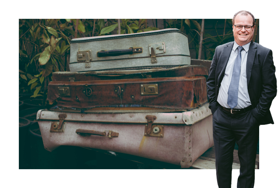 Pictured; Adam Wightman, head of Canny Legal, standing and smiling in a black and grey suit with his hands in his pockets. Behind him is an image of three old and worn suitcases stacked on top of one another.