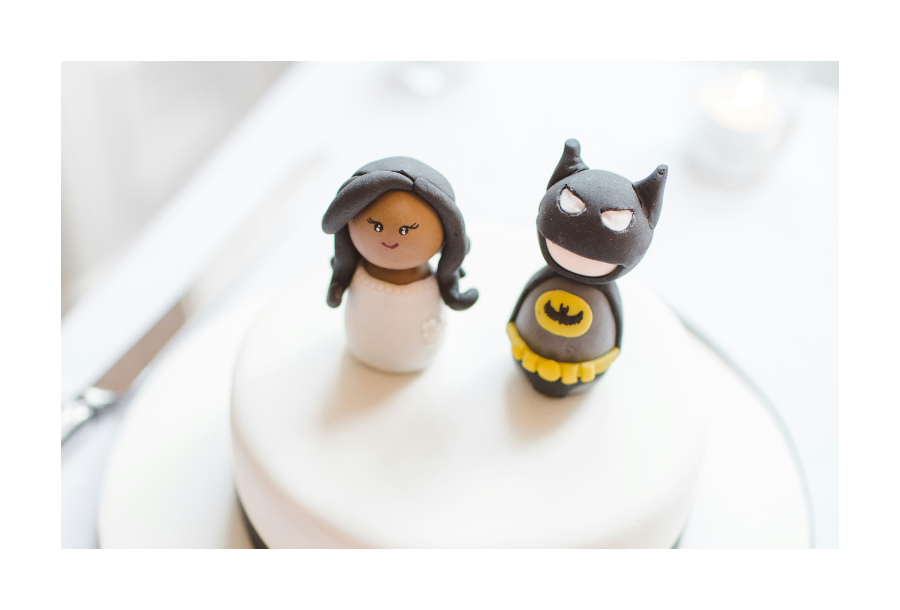 Pictured, a white muted background of a table, in the foreground there is a white frosted wedding cake. On the top of the cake are two figurines, the male represented as a batman figure and the female in a white dress and black long hair.