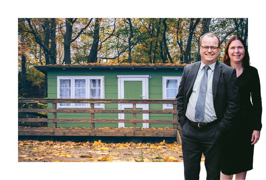 Pictured: Adam and Karlene Wightman standing smiling in business attire. Behind them is an image of a small green house covered in autumn leaves.