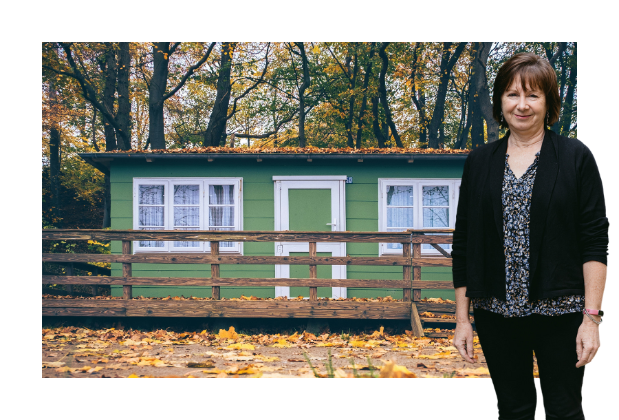 Pictured: Nancy Robertson standing smiling with her hands by her side, wearing a floral top with black pants and black cardigan. Behind her is a picture of a green house with lots of fallen leaves.