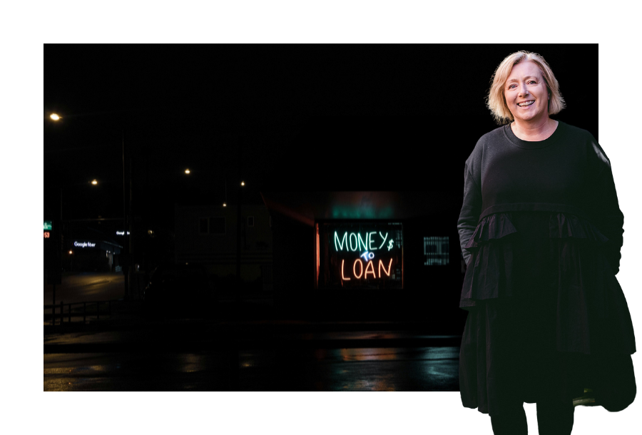 Pictured, Krystine Canny-Smith wearing a long sleeve black dress and black stockings, her hands are in her dress pockets. In the background is a dark street with a sign that is lit up and says "Money to Loan" ion neon colours.