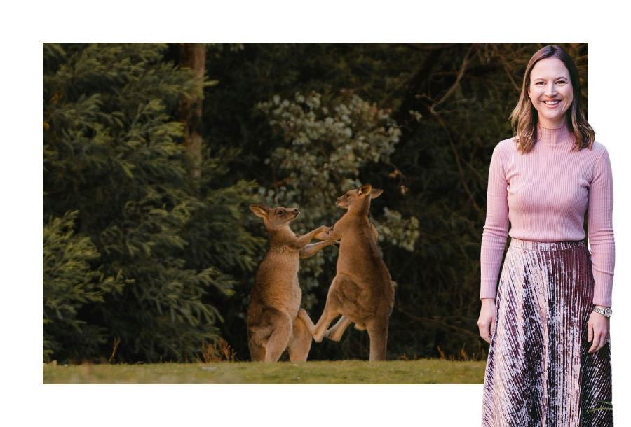 Pictured: Gabrielle Andersen standing smiling wearing a pink long sleeve top and a pink long skirt. Behind her is an image of two kangaroos fighting.