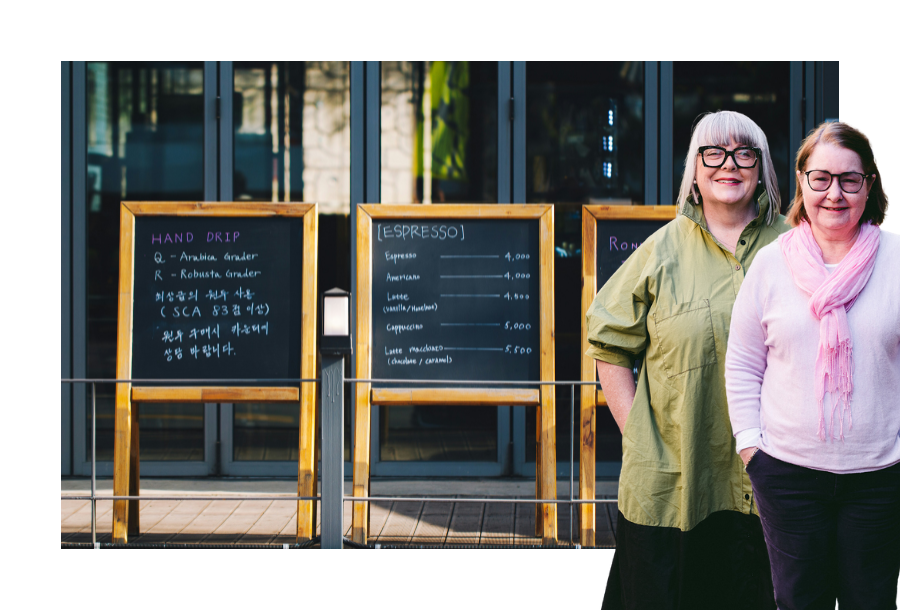 Pictured: Anthea Taylor and Christine Friel standing smiling. Behind them is an image of three chalk menu boards out the front of a cafe.