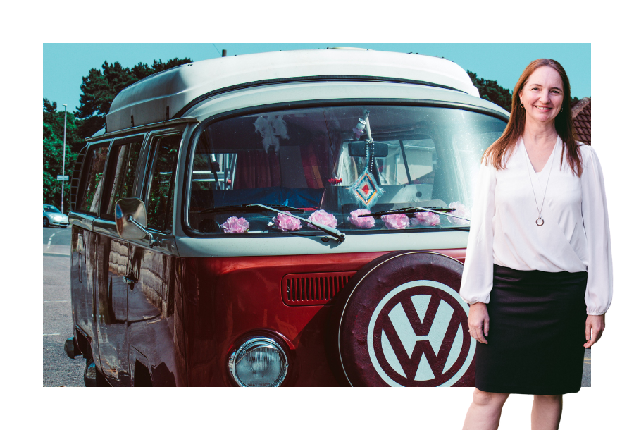 Pictured: Samantha Butcher standing smiling in a long sleeve white top and black skirt. Behind her is an image of a white and red Van with pink flowers on the dashbaord.