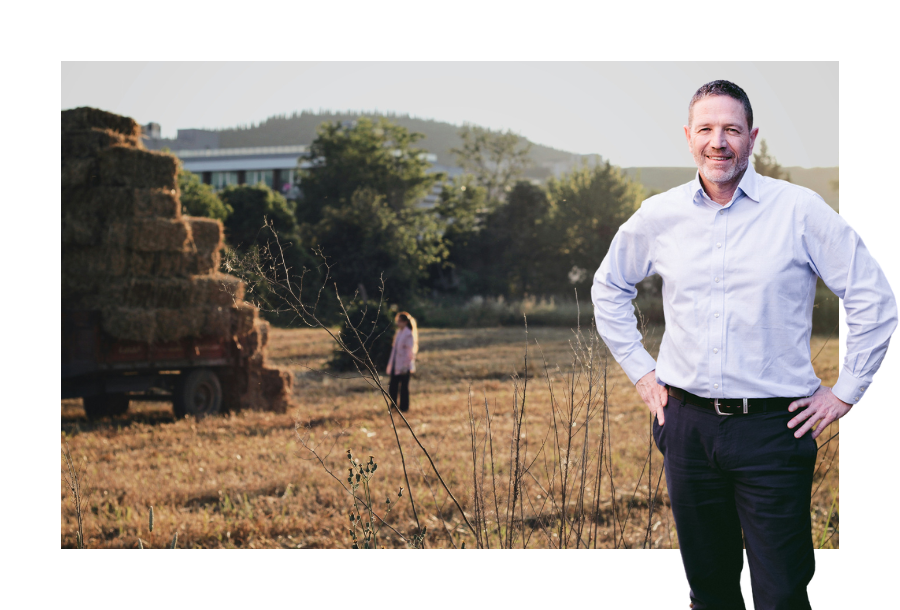 Pictured: Adam Ramage standing and smiling in a powder blue business shirt and black trousers. Behind him is an image of a tractor load of hay bales with a women standing behind.