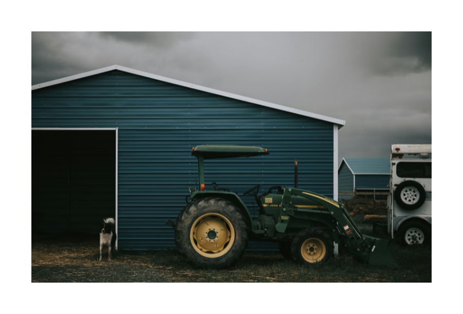 Pictured, a dark and gloomy outside setting. You can see a green John Deere tractor sitting outside a dark blue coloured farm shed. On the left hand side is a gorgeous black and white farm dog.