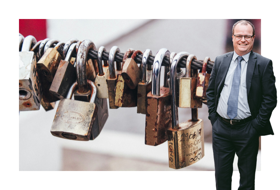 Pictured: Adam Wightman standing smiling in a dark grey suit with his hands in his pockets. Behind him is an image of many padlocks locked onto a thin pole.