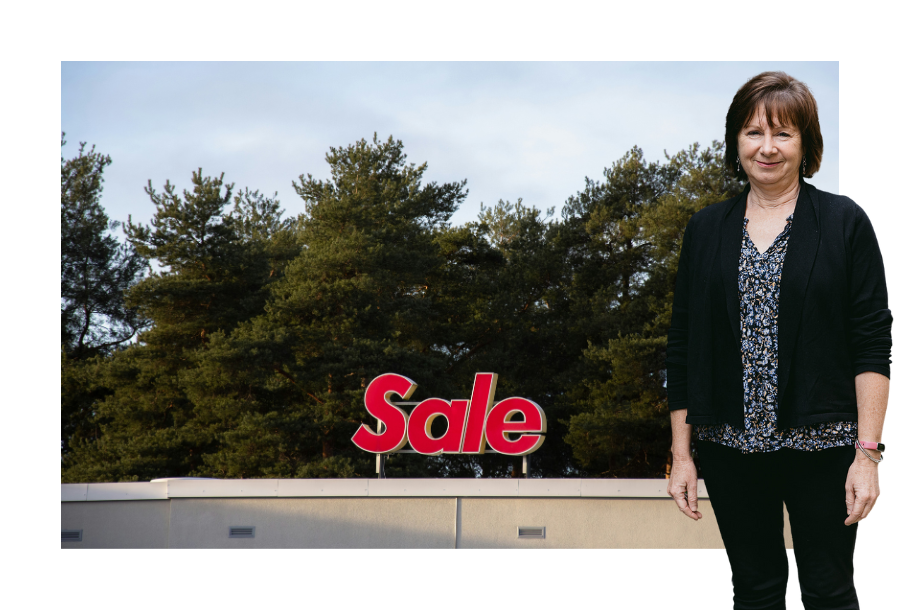 Pictured: Nancy Robertson standing smiling with black pants, a floral blue shirt and a black cardigan. Behind her is an image of a "Sale" sign on top of a building with treetops in the background.