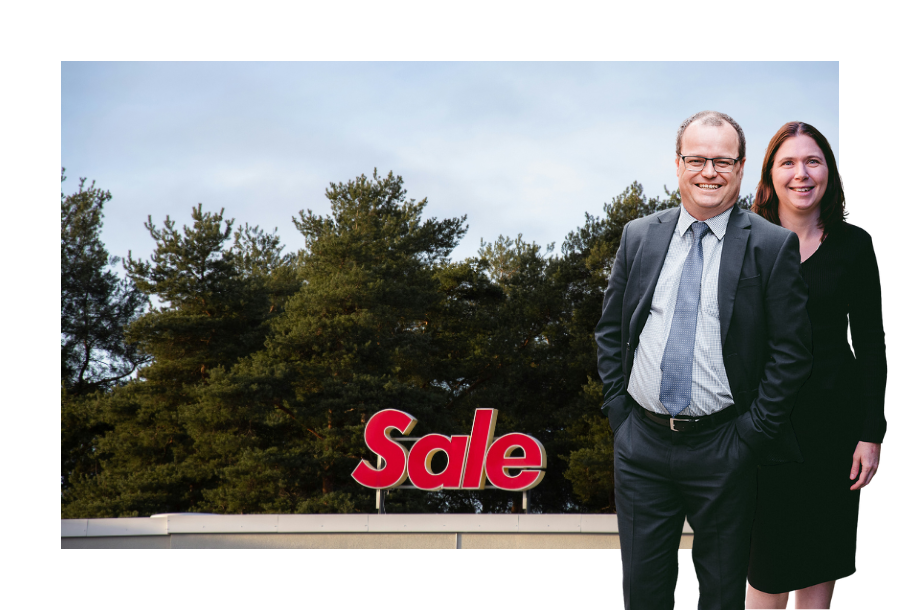 Pictured: Adam and Karlene Wightman standing smiling in business attire. Behind them is an image of a red sign on top of a building with trees in the background, the sign reads; "Sale".