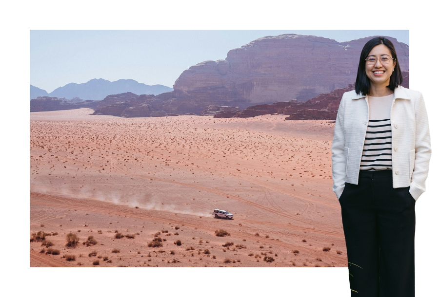 Pictured: Helen Yau standing smiling with her hand in her pockets, wearing a beige cardigan and black trousers. Behind her is an image of a car driving across an arid desert landscape with Rocky Mountains in the background.