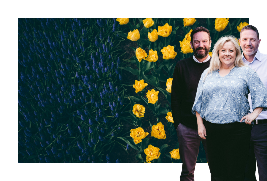 Pictured: Canny Accounting Managers, Danny, Michelle and Adam standing smiling. In the background is an aerial view of a field of flowers. On the left are deep purple lillies and on the right hand side is bright yellow daffodils.