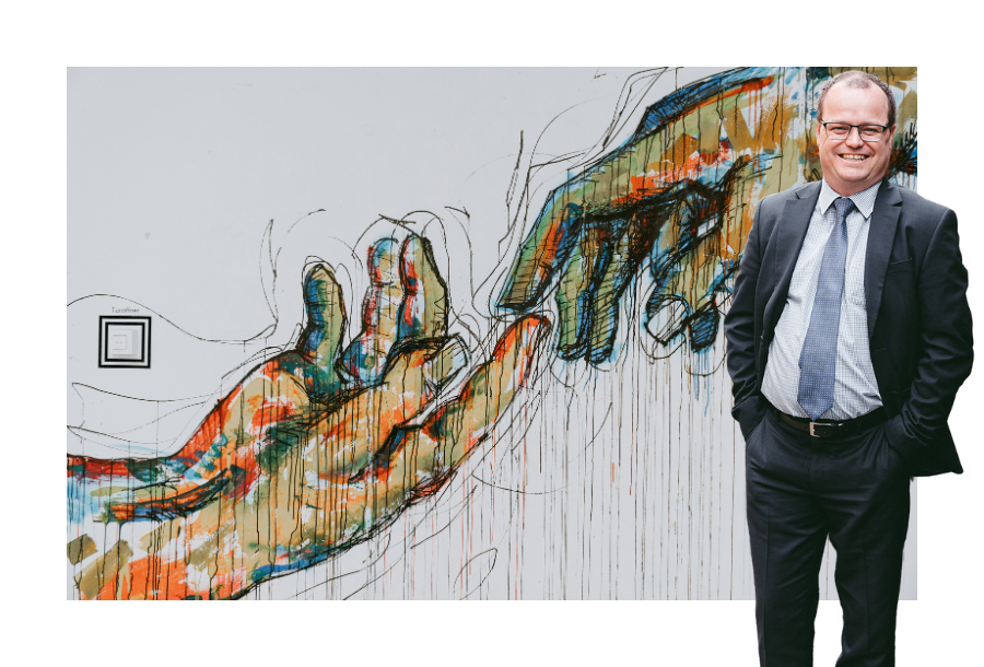 Pictured: Adam Wightman standing smiling wearing a black suit with a light blue shirt and blue tie with his hands in his trouser pockets. Behind him is an art piece painted on a wall depicting two hands with the pointer fingers touching.