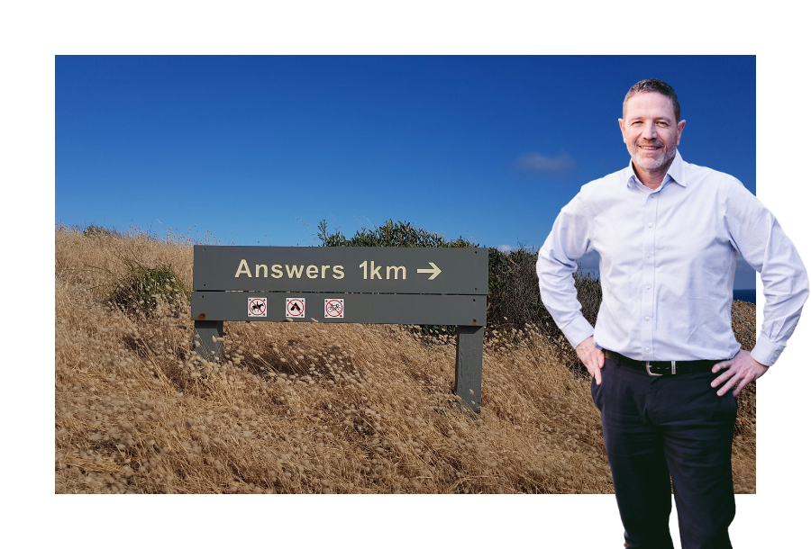 Pictured: Adam Ramage wearing a white shirt and black trousers, with his hands on his hips. Behind him is an image of a dark green painted wooden sign that reads "Answers, 1km" with an arrow pointing to the right.